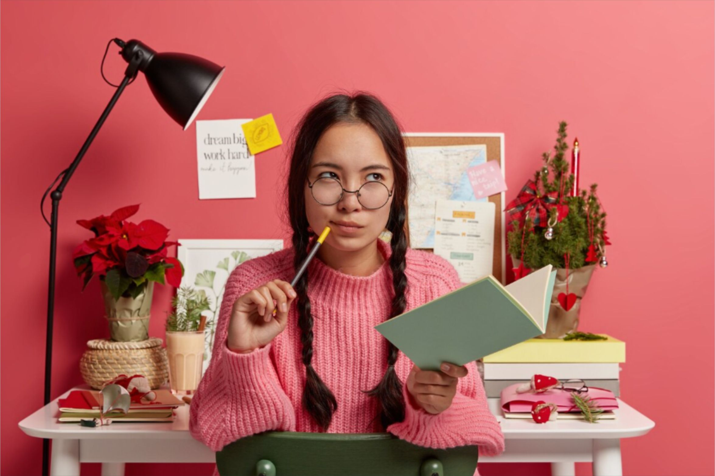Seorang perempuan muda berambut kepang sedang berpikir ide bisnis online modal kecil di meja studinya, memegang pulpen di dekat bibirnya, dengan buku terbuka dan laptop di hadapannya, serta lampu belajar dan tanaman di latar belakang berwarna merah muda.